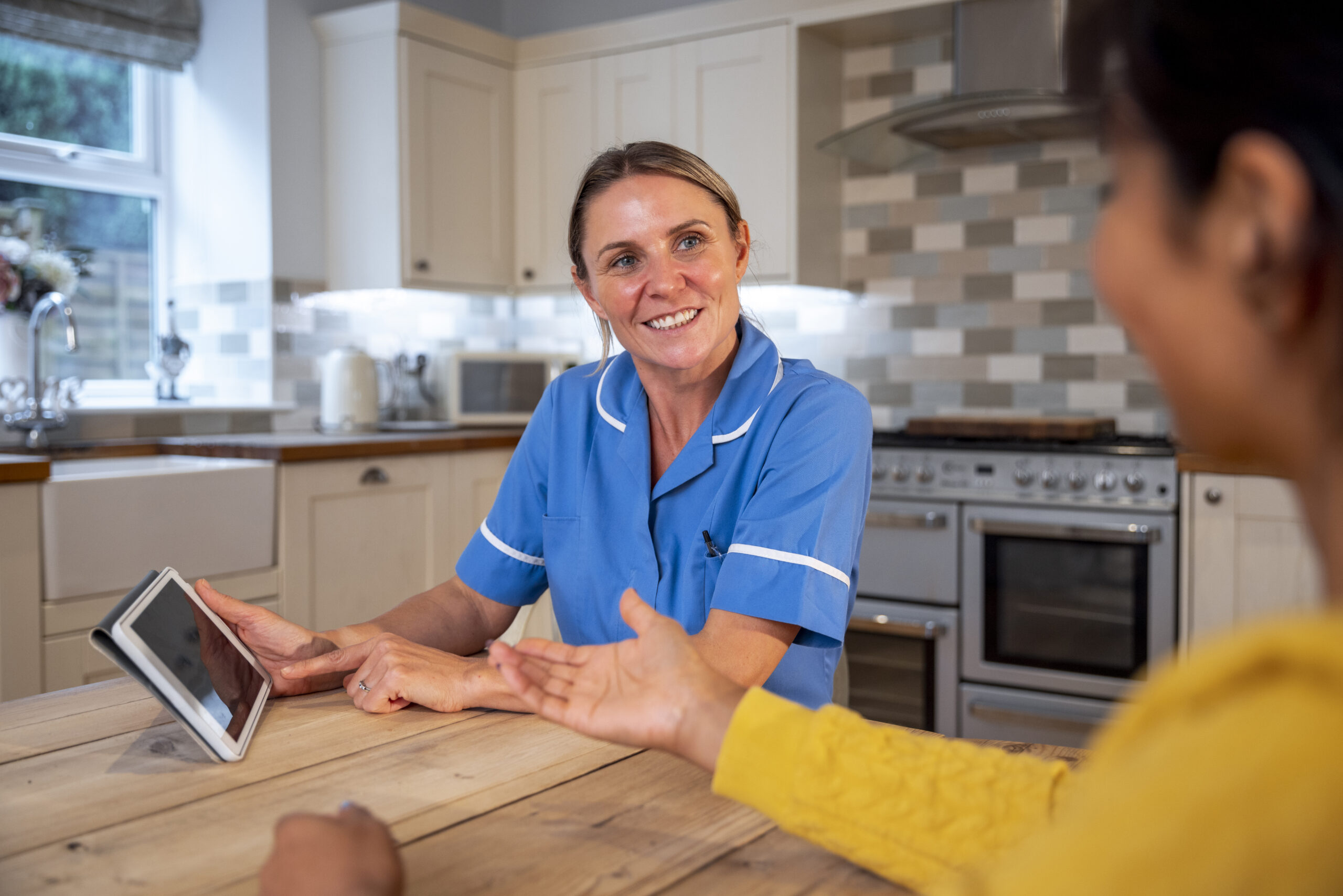 A woman talking to an on call nurse who has come to visit her at home, they are both sitting at the dining table in the kitchen. The nurse is talking to the patient and using a digital tablet as an aid.