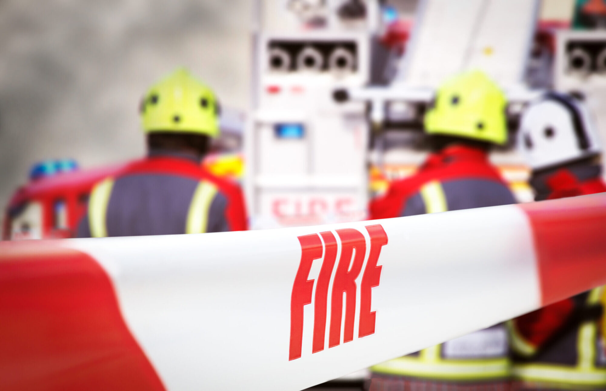 Scene of incident is cordoned off with red and white tape, reading ‘fire’. Out of focus in the background are three firefighters, two fire engine appliances and smoke billowing through the air.