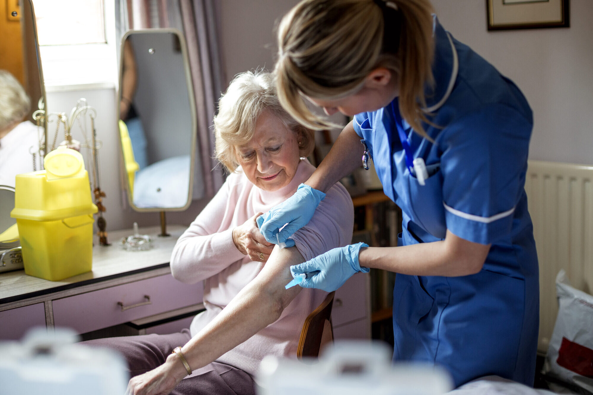 A nurse visits an elderly woman in her home. The elderly woman is happily sitting on a chair at her dressing table.